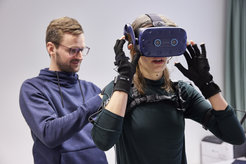 Florian Binder befestigt Sensoren am Körper seiner Kollegin Dorothee Pöhlchen, die ihre Bewegungen messen und in die virtuelle Realität übertragen. Eine Virtual-Reality-Brille versetzt sie in eine von Binder programmierte Umgebung.